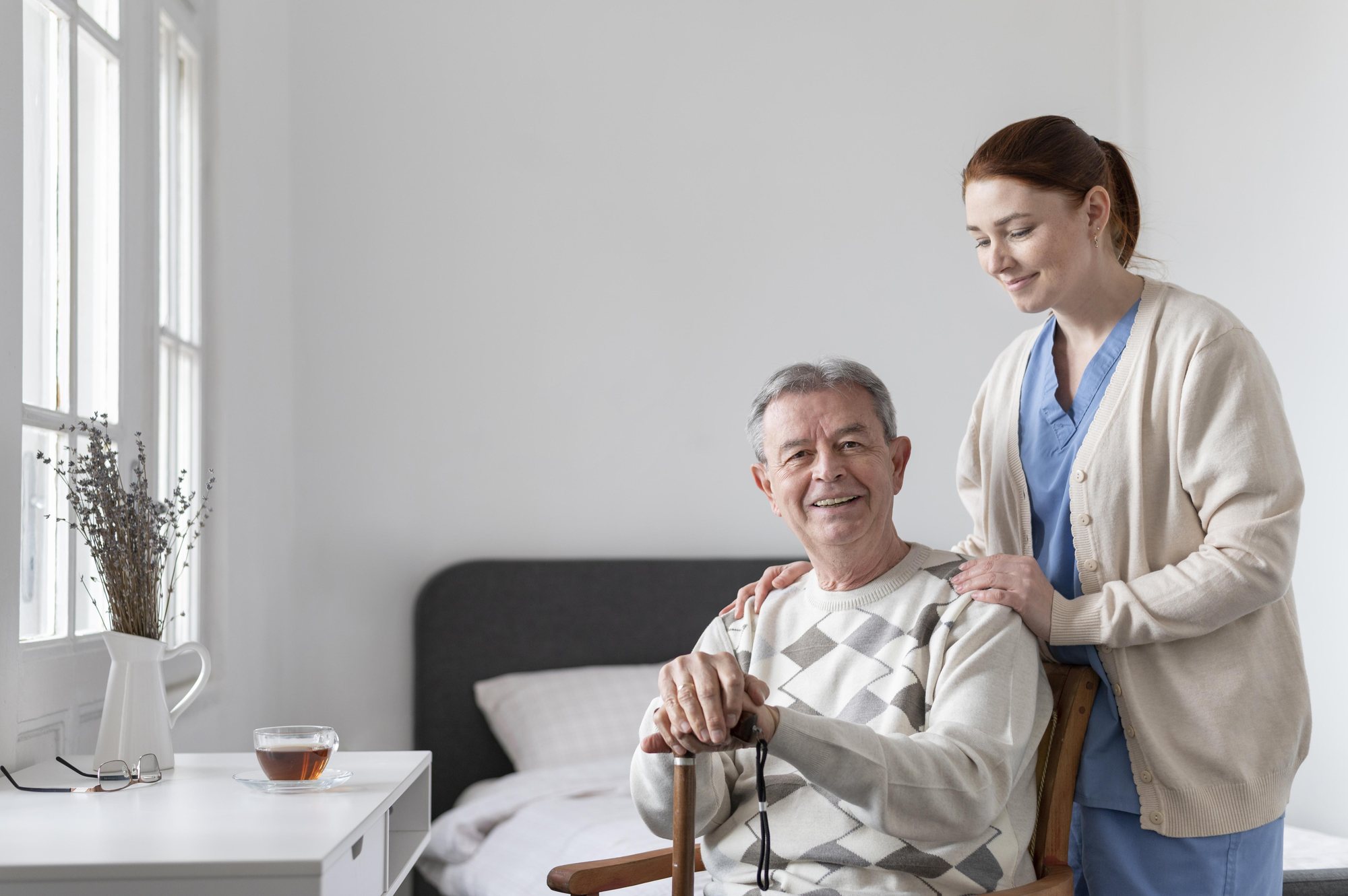 Cuidadora com as mãos nos ombros de um senhor sorridente sentado no quarto